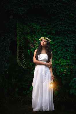 photo of woman in white dress and flower crown carrying small lantern while standing near vine plant